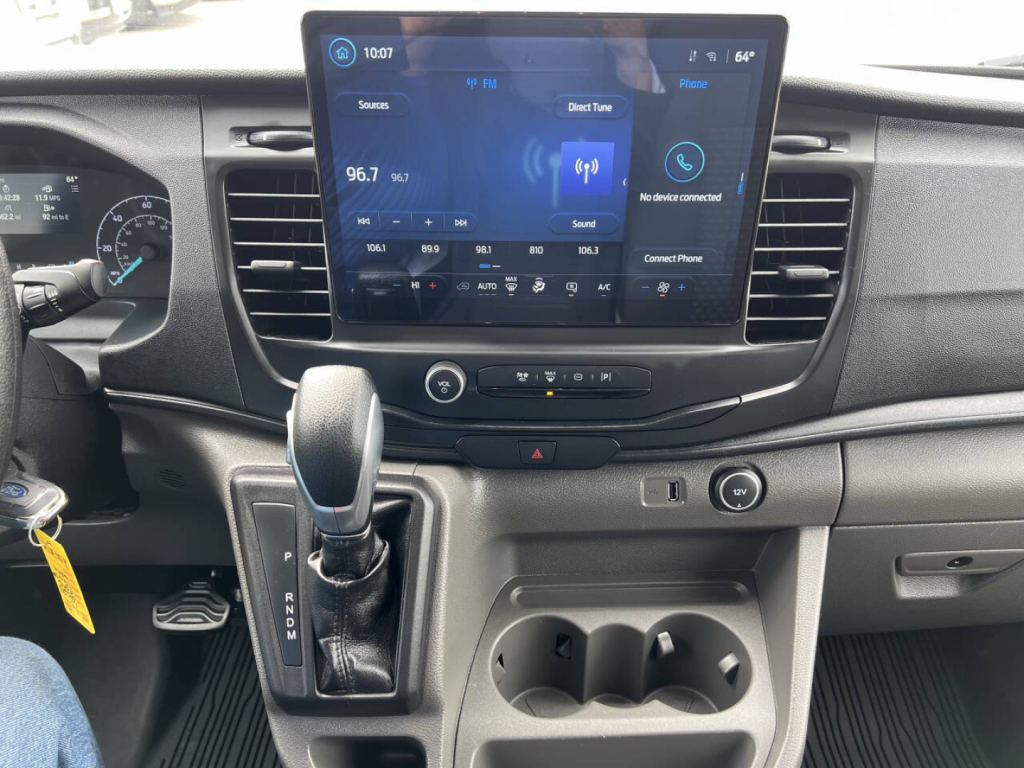 Interior of a Ford Transit van showing a center touchscreen infotainment display, gear selector, cupholders, and dashboard controls.