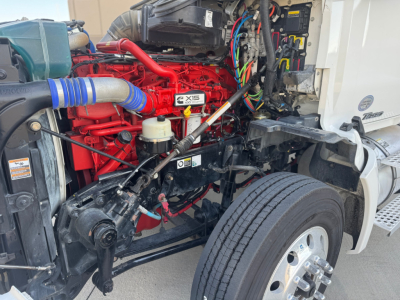 Kenworth T680 engine bay showing red X15 Cummins engine with blue intake hose and exposed wiring under hood; front wheel and chassis visible