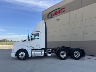 Kenworth T680 tractor-trailer in front of a building with MHC logo. Side view showing cab, fuel tanks, and dual rear axles.