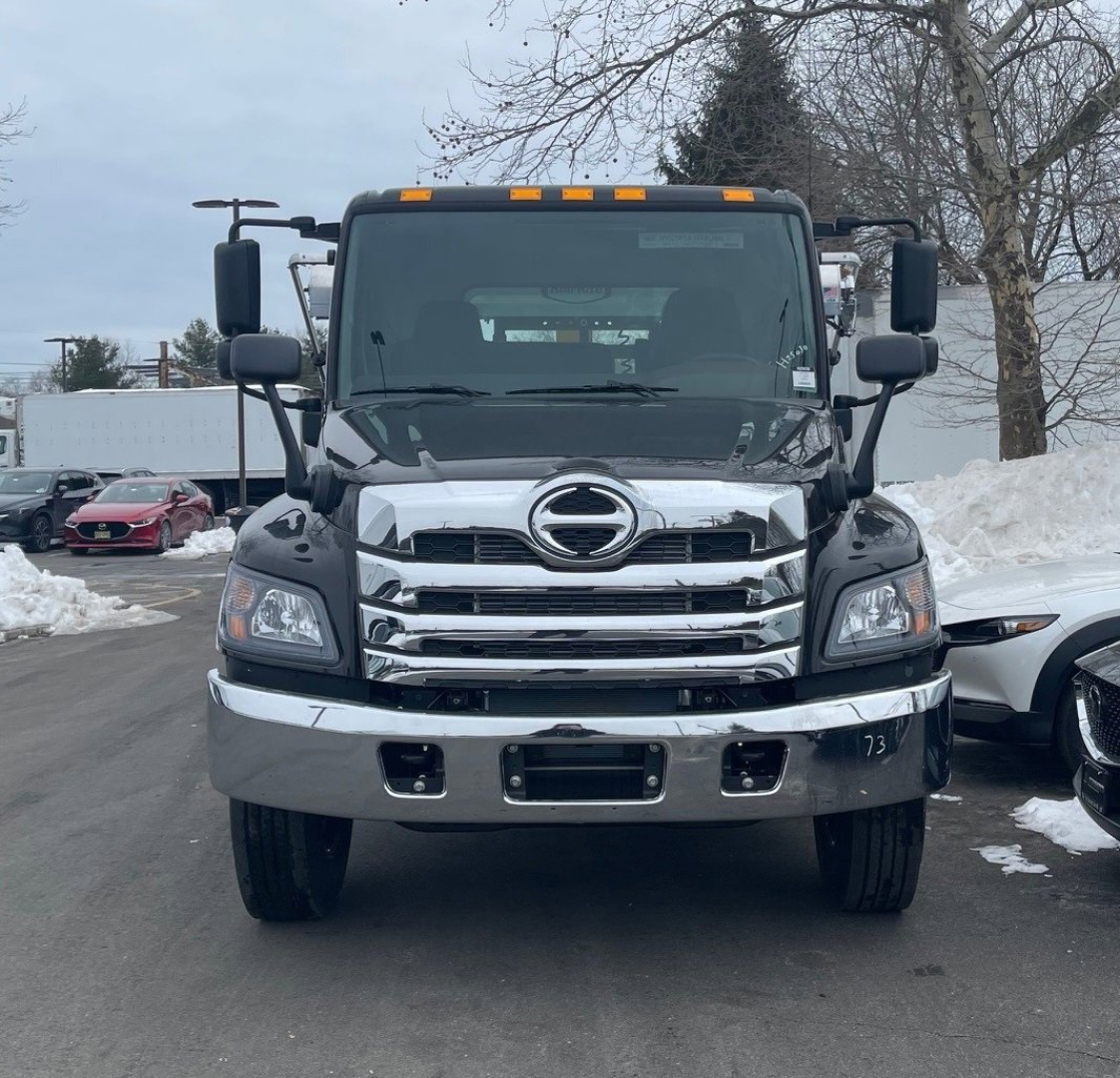 Front view of a 2026 Hino L7 hooklift truck showing chrome grille and bumper, Hino emblem, large headlights, and side mirrors.