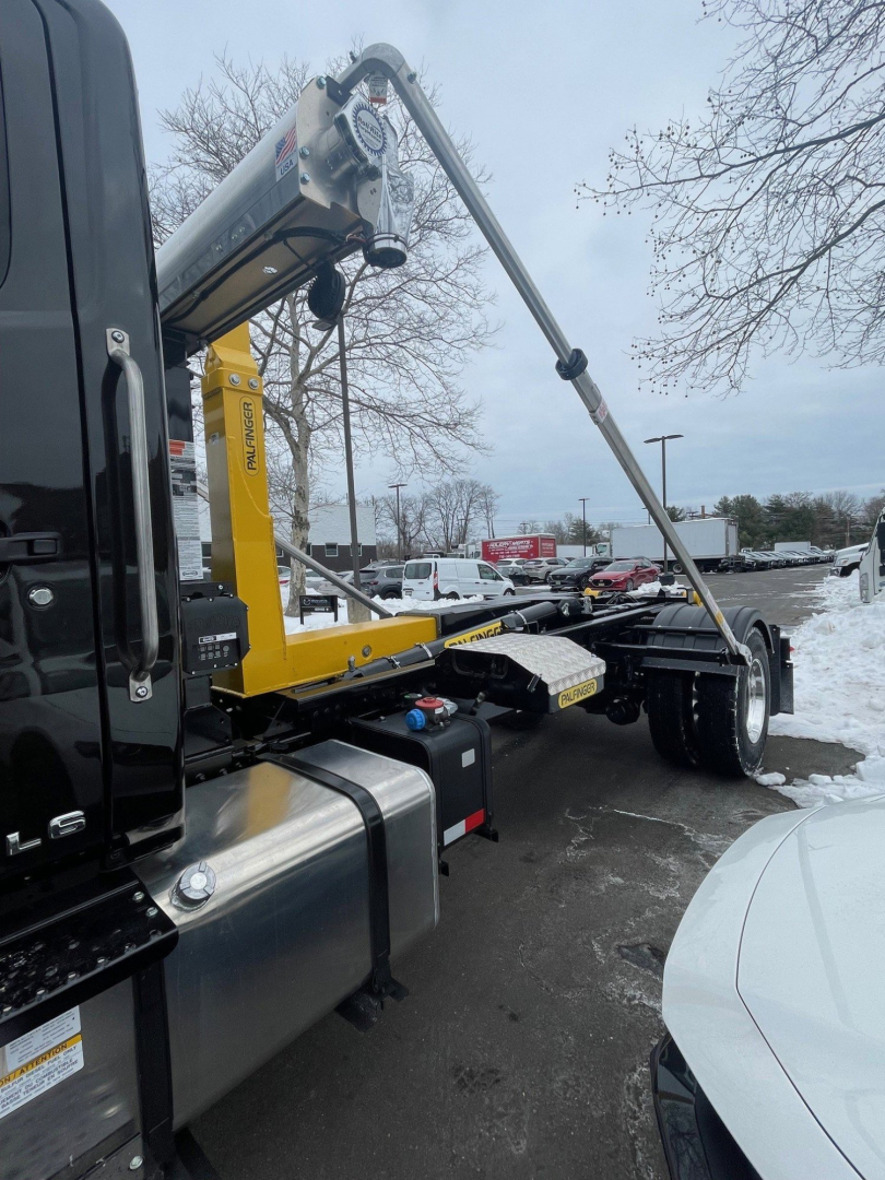 Truck chassis with a yellow hooklift crane arm extended, mounted on a black cab and dual rear wheels visible, showing a hydraulic lifting mechanism and metal frame.