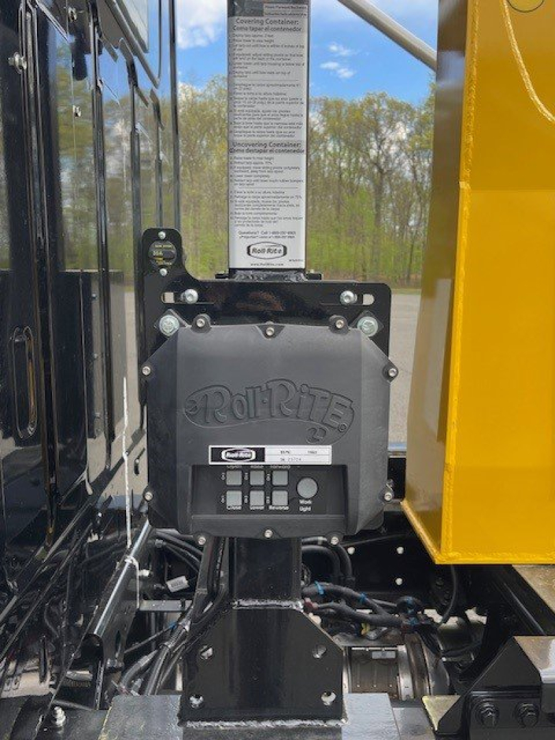 Truck component showing a Roll-Rite hoist control box mounted on a black frame with hydraulic lines and a yellow body side panel visible in bright daylight.