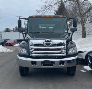 Front view of a 2026 Hino L7 hooklift truck showing chrome grille and bumper, Hino emblem, large headlights, and side mirrors.