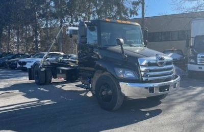 New 2026 Hino L7 hooklift truck front-left view showing cab, chrome grille, and hooklift behind cab.