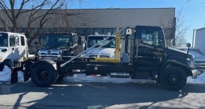 New 2026 Hino L7 hooklift truck side view showing cab, chassis, and hooklift arms with black paint and yellow lifting structure.