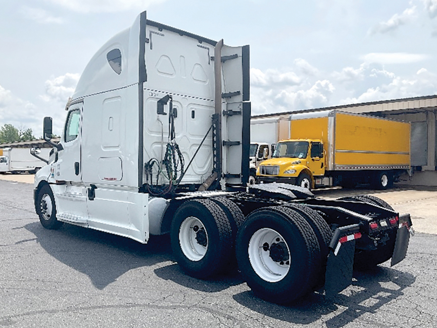 Truck rear view showing white Freightliner Cascadia126 sleeper cab with dual rear axles and fifth wheel; yellow box truck in background.
