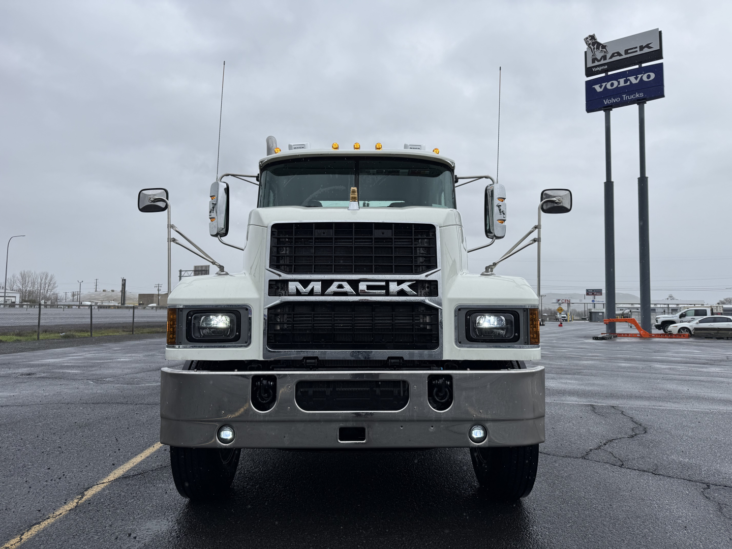 Truck front view of a white 2026 Mack PI64T with large grille, bumper, and dual square headlights; Mack logo on grille; dealership sign in background.