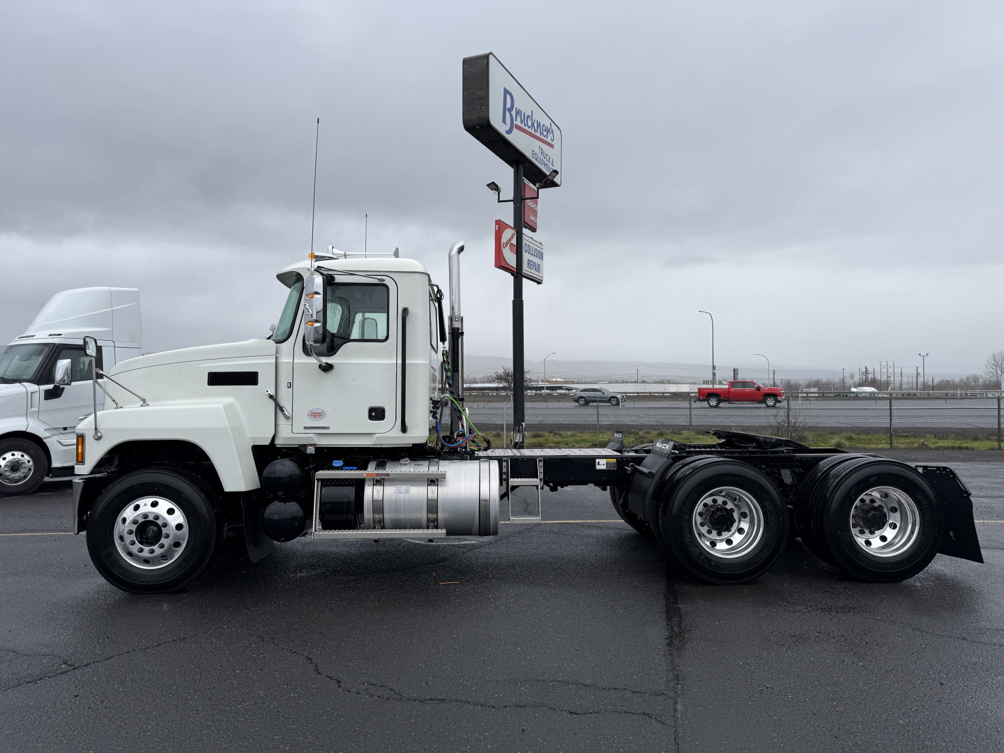 Side view of a white Mack PI64T truck chassis with cab, fuel tanks, and three rear axles on a blacktop lot.