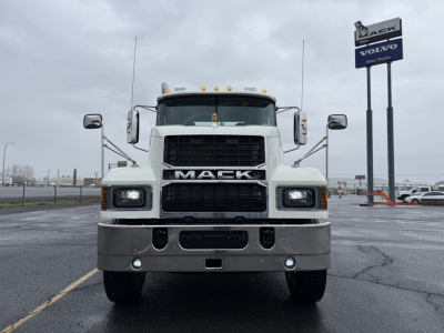 Truck front view of a white 2026 Mack PI64T with large grille, bumper, and dual square headlights; Mack logo on grille; dealership sign in background.