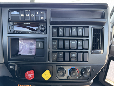 Dashboard area of a Mack truck cab showing gear selector R N D, radio, various switches, and climate controls. Visible text includes MACK on the radio, “R N D” letters, knobs, and a red trailer brake knob.