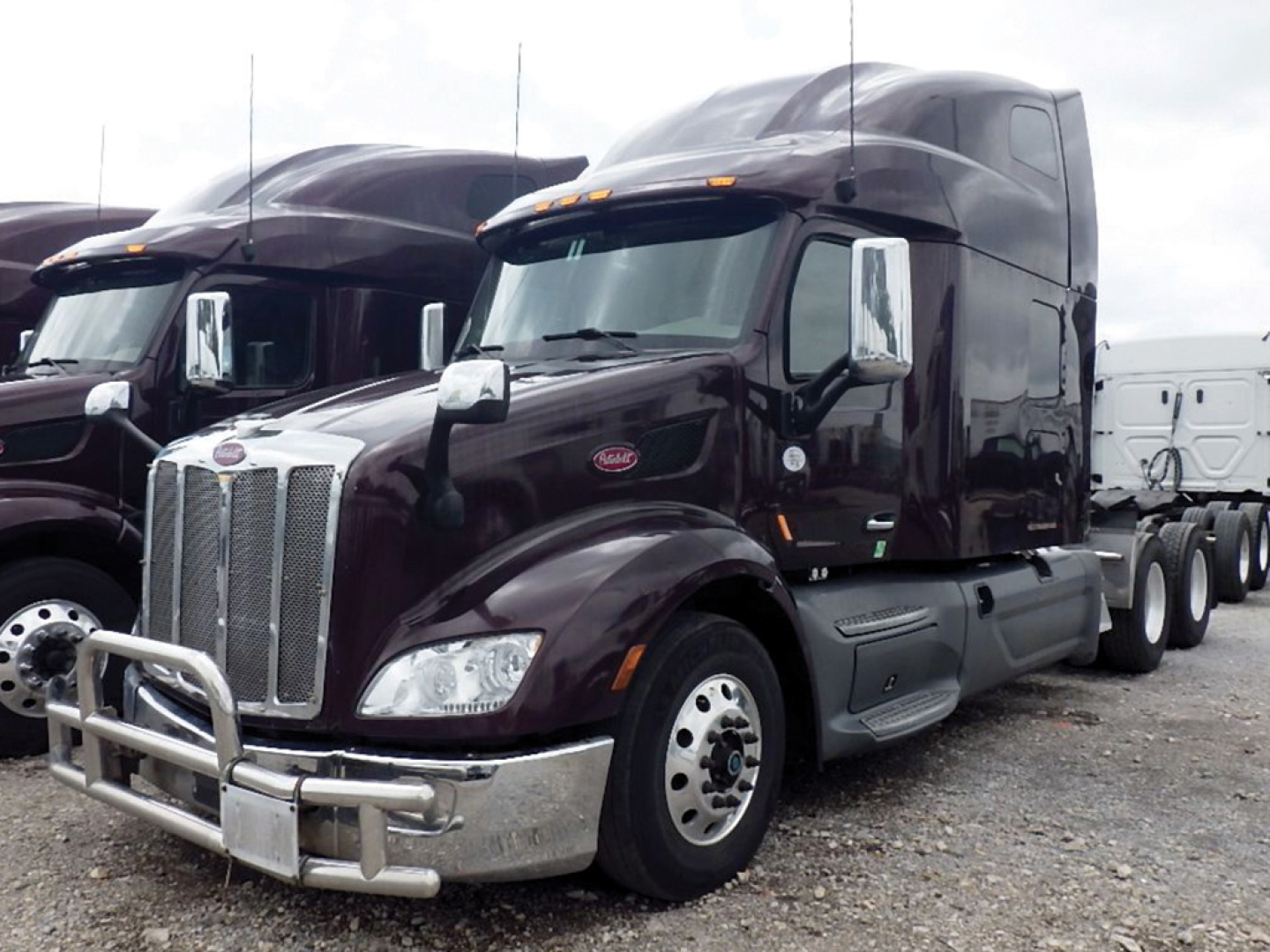 Peterbilt 579 truck in dark maroon with chrome bumper guard and large grille, sleeper cab, side mirrors, visible front wheel, trailer connection in background.