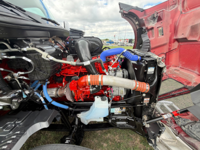 Truck engine bay of a 2026 Kenworth T680 with exposed red engine and blue hoses, hood open, various components visible.