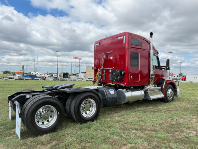 Kenworth T680 conventional sleeper truck, red cab, rear view showing dual rear axles, fuel tanks, and fifth wheel, highway yard in background.