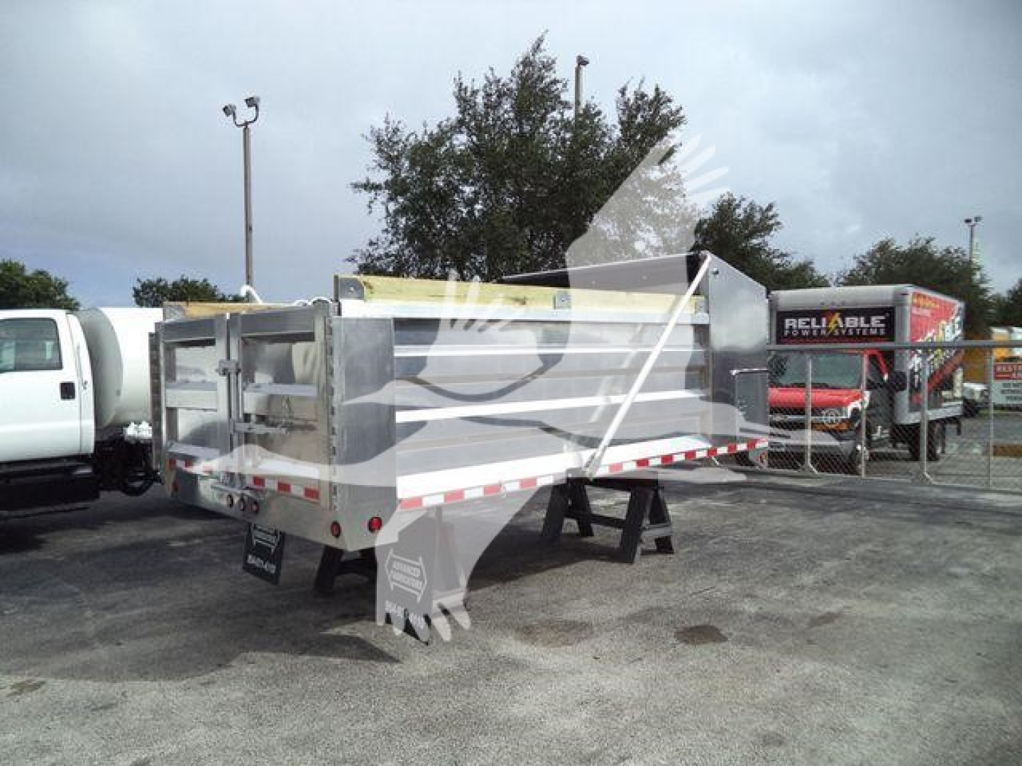 Dump trailer showing stamped aluminum bed with side rails and rear doors; truck yard with other equipment in background.
