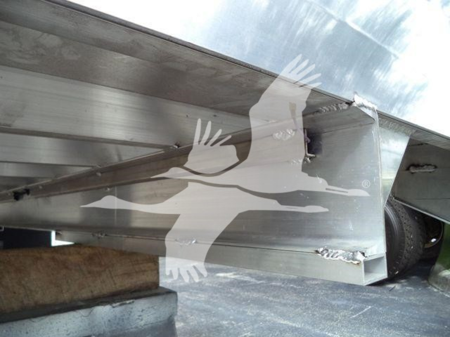 Truck dump trailer undercarriage showing metal frame and welded corner with a tire in background; faint watermark graphic of a flying bird overlaying the image.