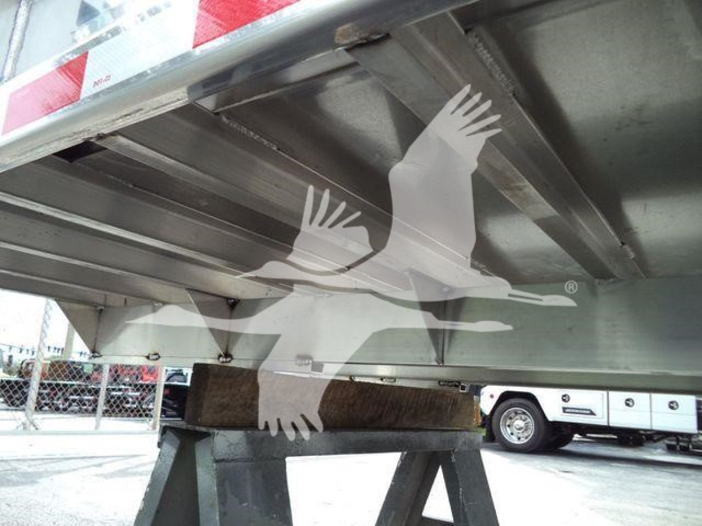 Dump truck bed underside showing structural beams with a wooden block support and wheel in background.