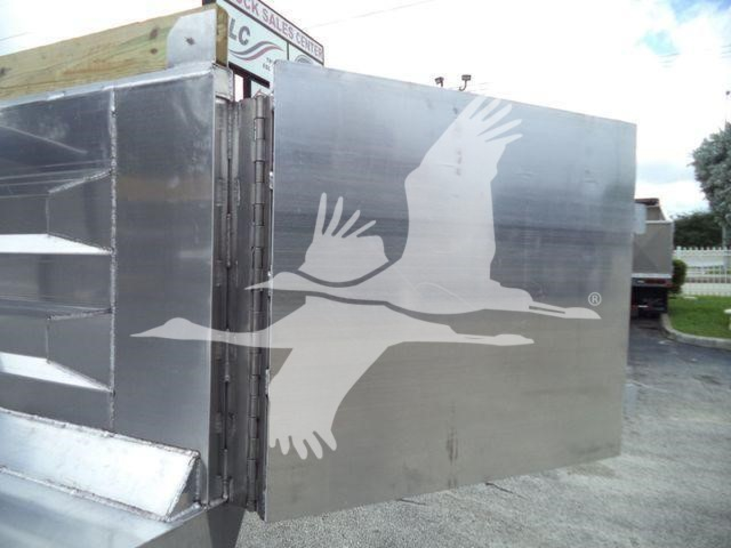 Dump truck body with metal panel showing a white bird graphic in flight. Side view reveals a dump bed with hinges and support, aluminum finish.