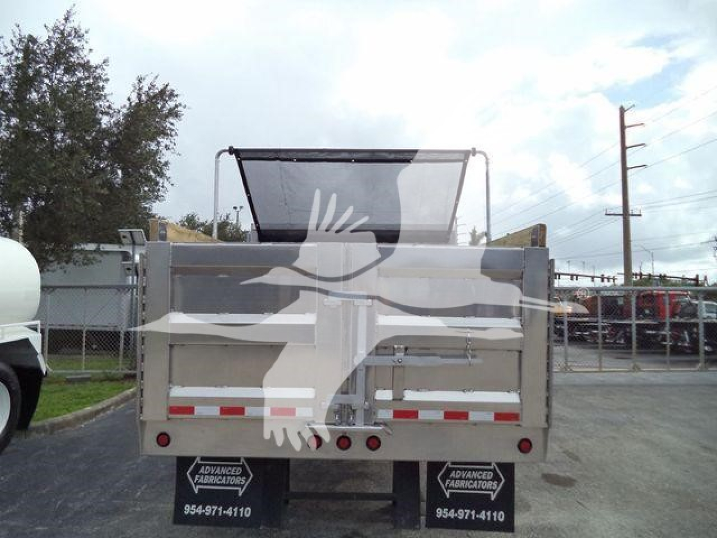 Rear view of a 14 ft dump bed truck attachment in aluminum with high side walls, hydraulic lift mechanism open tailgate, road trailers and lot in background.
