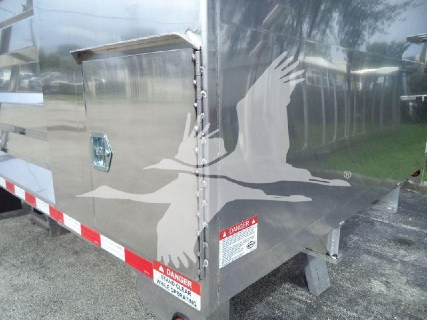Aluminum dump trailer side showing bird graphic, access door, warning labels, and reflective safety strip along bottom edge.
