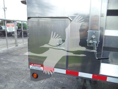 Back of 14 ft dump trailer with metal exterior, safety latch, red/white reflective tape, and warning decal near bottom left read 'DANGER STAND CLEAR WHILE OPERATING'