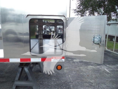Truck dump trailer side with open rear door showing interior and safety labels and a stand. Visible text includes 'DANGER STAND CLEAR WHILE OPERATING' sticker and reflection of equipment.