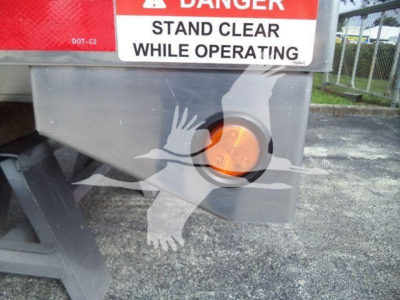 Dump bed side with safety labels: 'DANGER STAND CLEAR WHILE OPERATING' and an orange circular valve cap; partial logo silhouettes overlay.