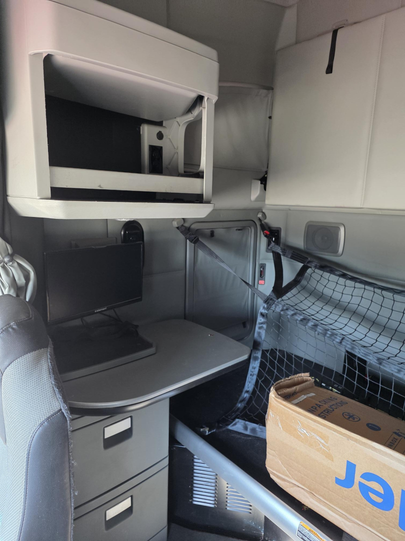 Interior of a Kenworth truck cab showing a storage shelf, overhead console, netting on the side, and a drawer cabinet.
