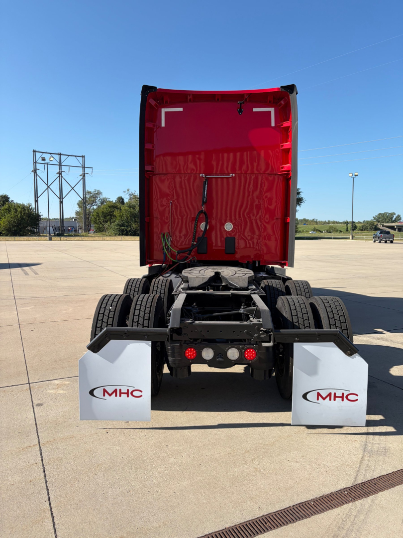 Kenworth truck rear with open red hood, dual rear axles, white mud flaps with MHC logo, in a parking lot.