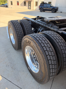 Dual rear truck wheels with heavy-duty tires on a Kenworth tractor chassis, frame and suspension visible, with other vehicles in background.