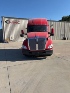Kenworth T680 front view in red with large chrome grille, headlights, and side mirrors; truck is in a parking lot with a metal warehouse background.