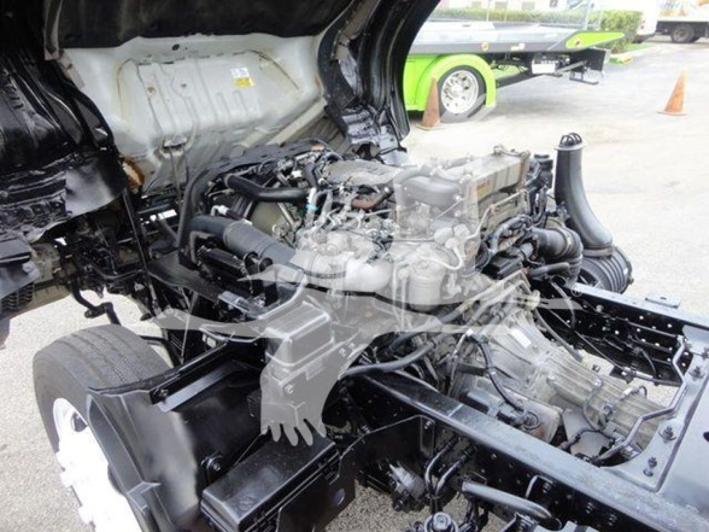Isuzu NQR engine bay view showing diesel engine and surrounding components; visible belts, hoses, and chassis frame; partial cab interior roof and firewall; utility truck undercarriage.