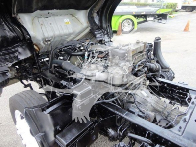 Isuzu NQR engine bay view showing diesel engine and surrounding components; visible belts, hoses, and chassis frame; partial cab interior roof and firewall; utility truck undercarriage.