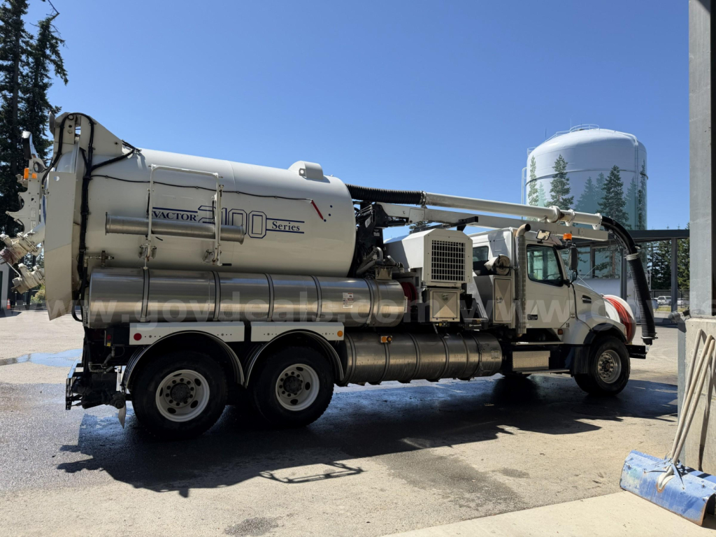 Volvo VHD vacuum truck with large cylindrical tank labeled Mac? 100 Series, hoses and pump assembly along side, industrial setting.