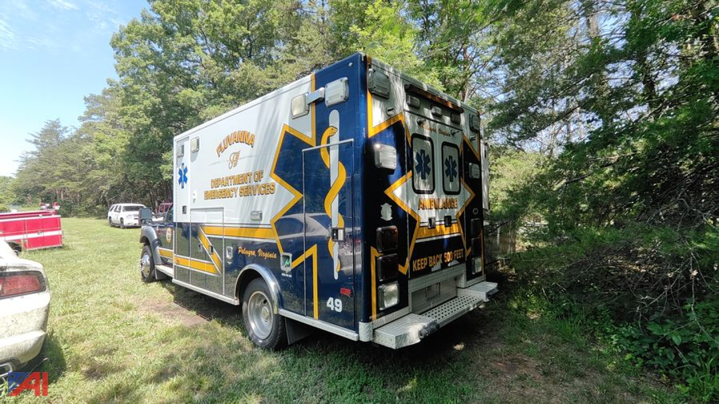 Ambulance body with blue, white and yellow graphics, rear doors, medical symbols, and text such as Ambulance, Emergency Medical Services, and 911; parked on grass with trees.