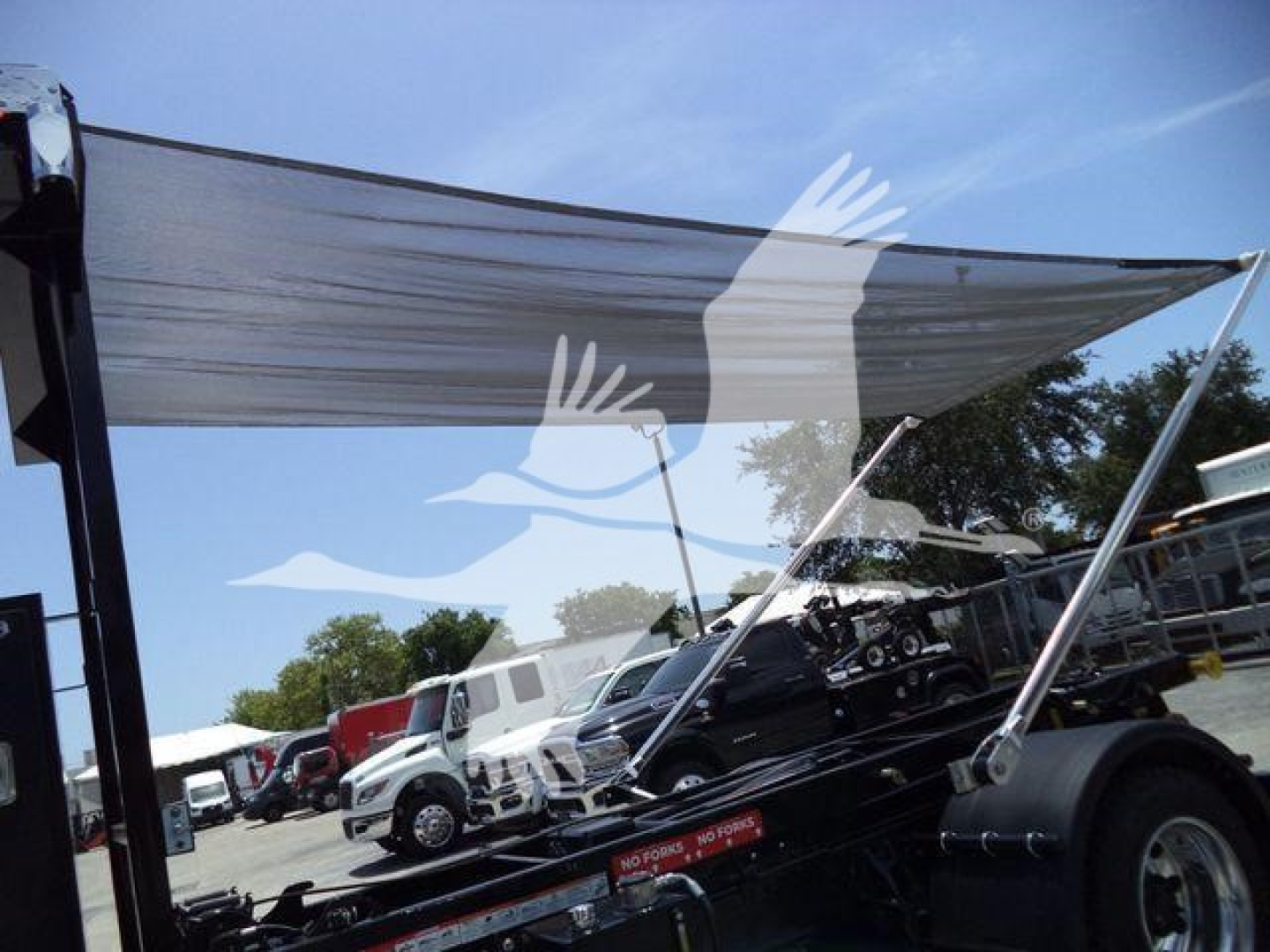 Truck trailer showing a raised shade sail and several trucks in a parking lot; no legible text visible on the image.