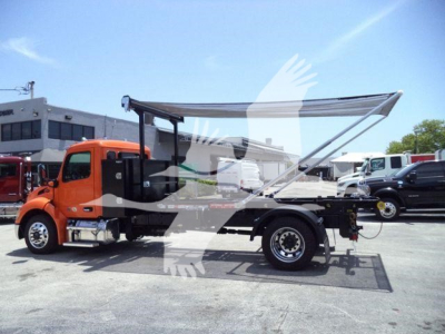Peterbilt 536 truck in a lot with a raised bed canopy and open rear storage; orange cab, black chassis, white trucks in background.