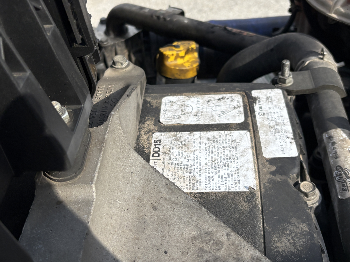 Truck engine bay showing a metal identification label and nearby yellow cap; partial view of hoses and components.