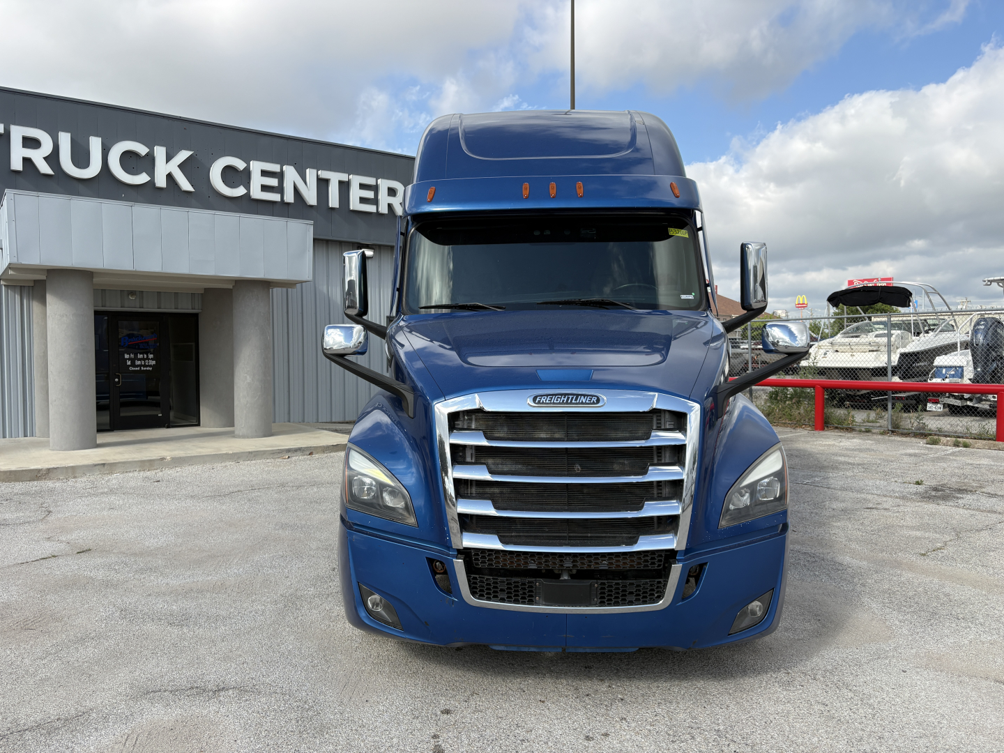Front view of a blue 2022 Freightliner Cascadia 126 tractor with chrome grille and cab mirrors, parked in lot in front of a truck center.