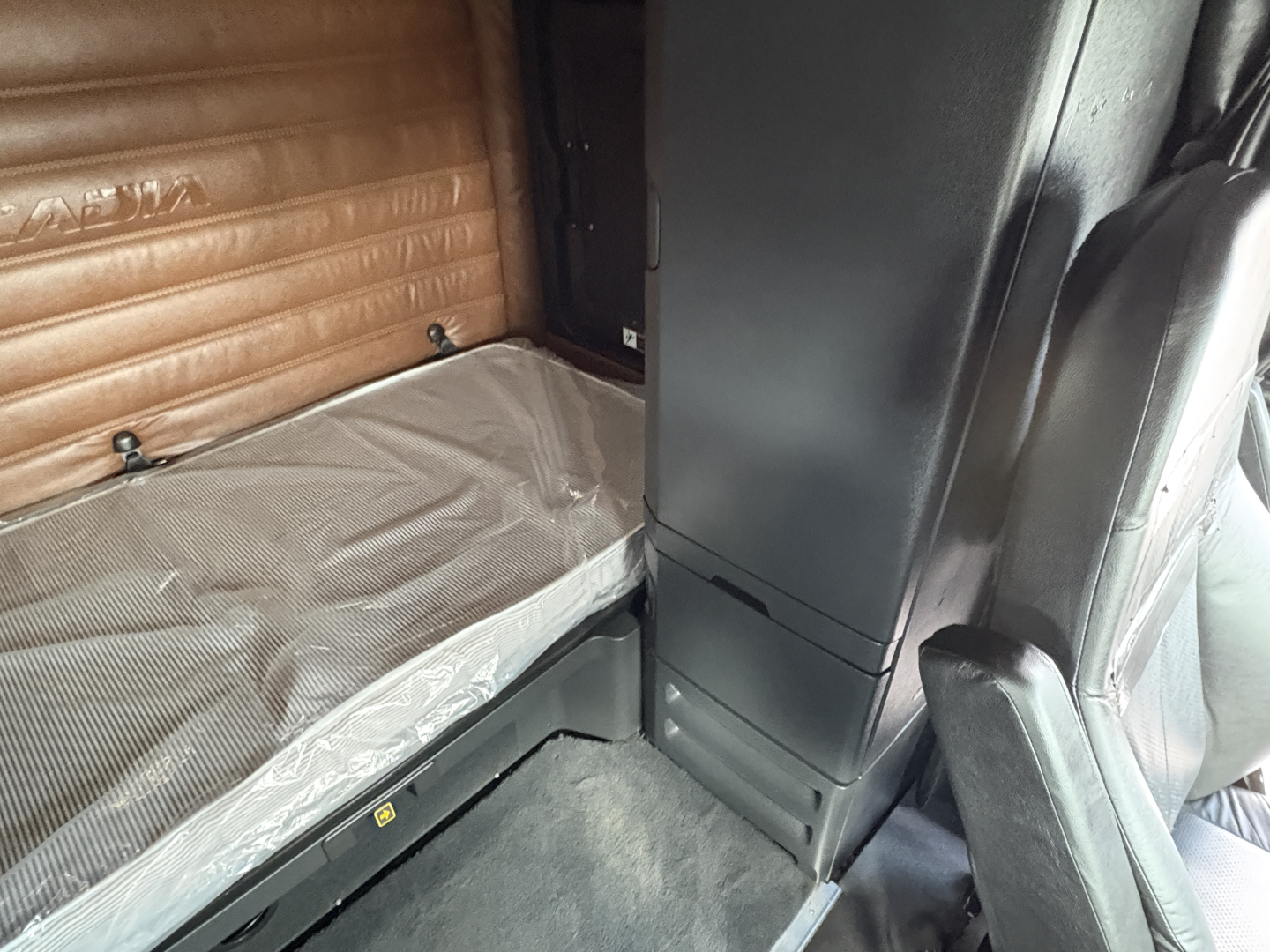 Cab interior of a 2022 Freightliner Cascadia 126 sleeper shows narrow bed with brown padded wall, vertical cabinet, and passenger seat; text not legible.
