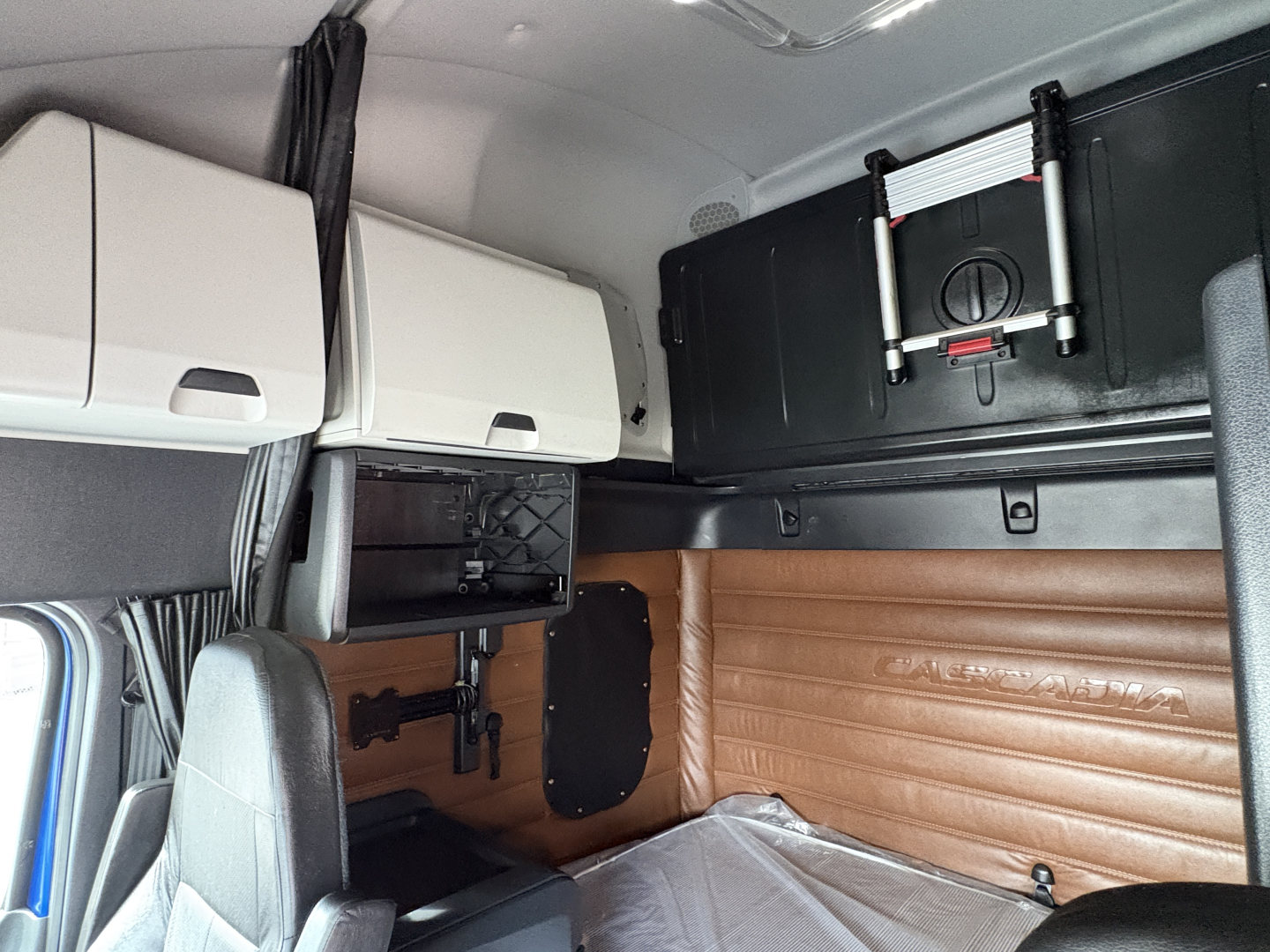 Interior of a Freightliner Cascadia cab showing overhead compartments, a mounted TV or monitor, and tan leather side paneling with a raised logo.