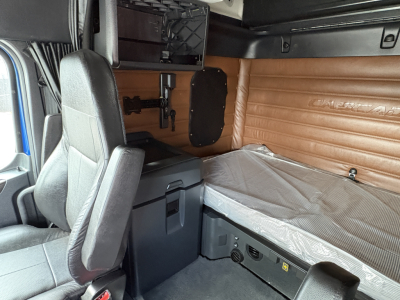 Truck cab interior of a Freightliner Cascadia 126 showing sleeper area with mattress, storage box, and seat. Visible details include tan side paneling and control panels.