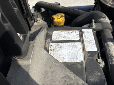 Truck engine bay showing a metal identification label and nearby yellow cap; partial view of hoses and components.