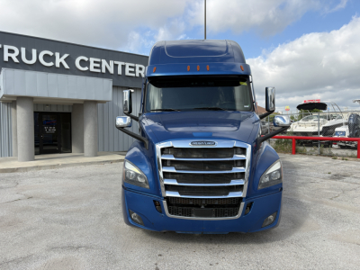 Front view of a blue 2022 Freightliner Cascadia 126 tractor with chrome grille and cab mirrors, parked in lot in front of a truck center.