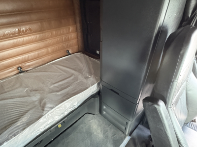 Cab interior of a 2022 Freightliner Cascadia 126 sleeper shows narrow bed with brown padded wall, vertical cabinet, and passenger seat; text not legible.