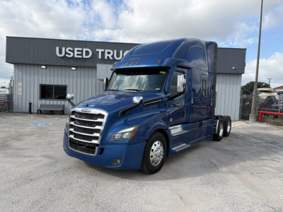 Freightliner Cascadia 126 sleeper tractor in blue parked outside a used truck lot, front three-quarter view showing chrome grille and cab details.