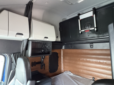 Interior of a Freightliner Cascadia cab showing overhead compartments, a mounted TV or monitor, and tan leather side paneling with a raised logo.