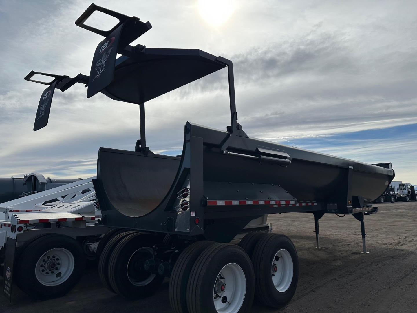 Frameless rock end dump trailer showing open-top bed and hydraulic lift, dual rear axles with white rims, black frame, and support legs.