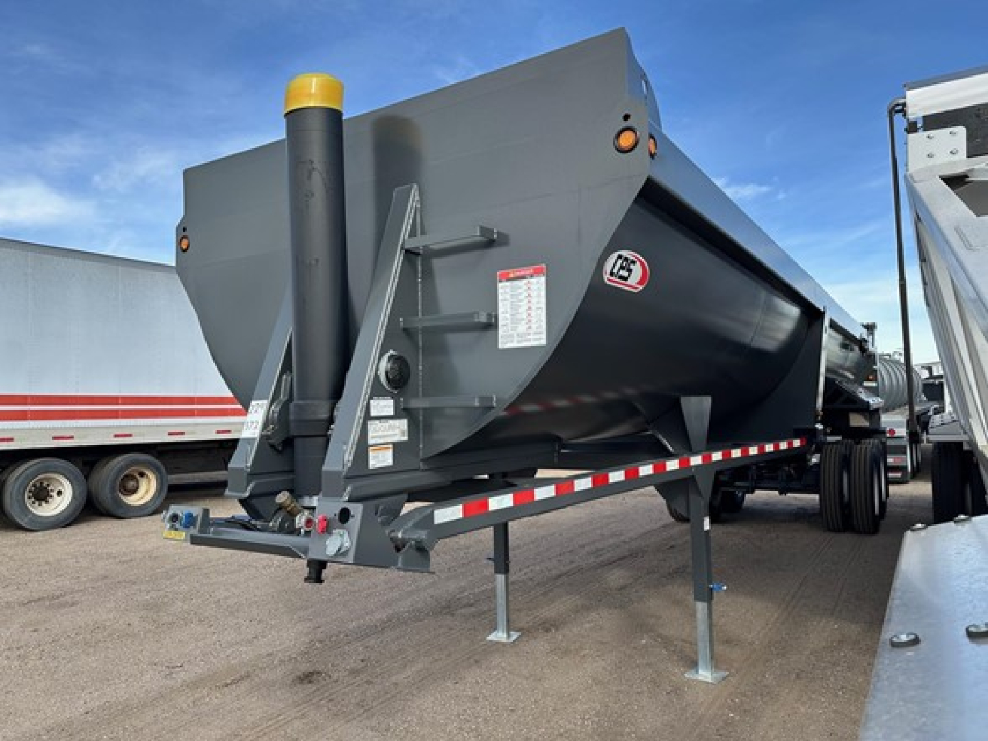 Frameless rock end dump trailer in a yard showing high lift gate and frame, with a yellow-topped chute and side access ladder; side markings and reflective strips visible.