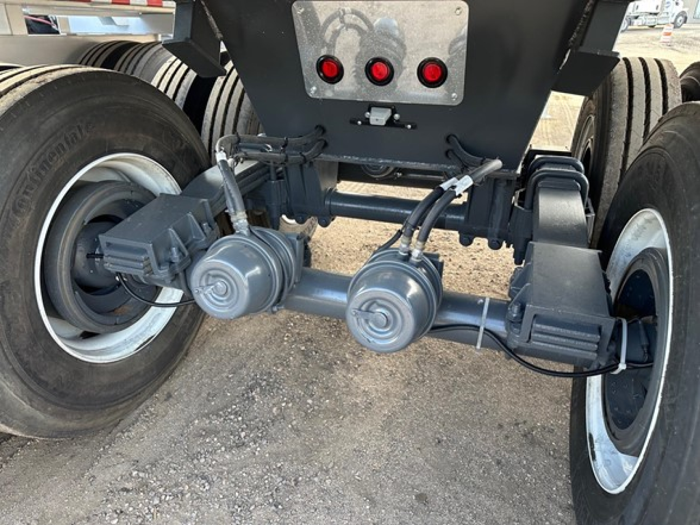 Trailer rear undercarriage showing dual axles with differential beams and suspension components, with red rear lights and a metal plate above.