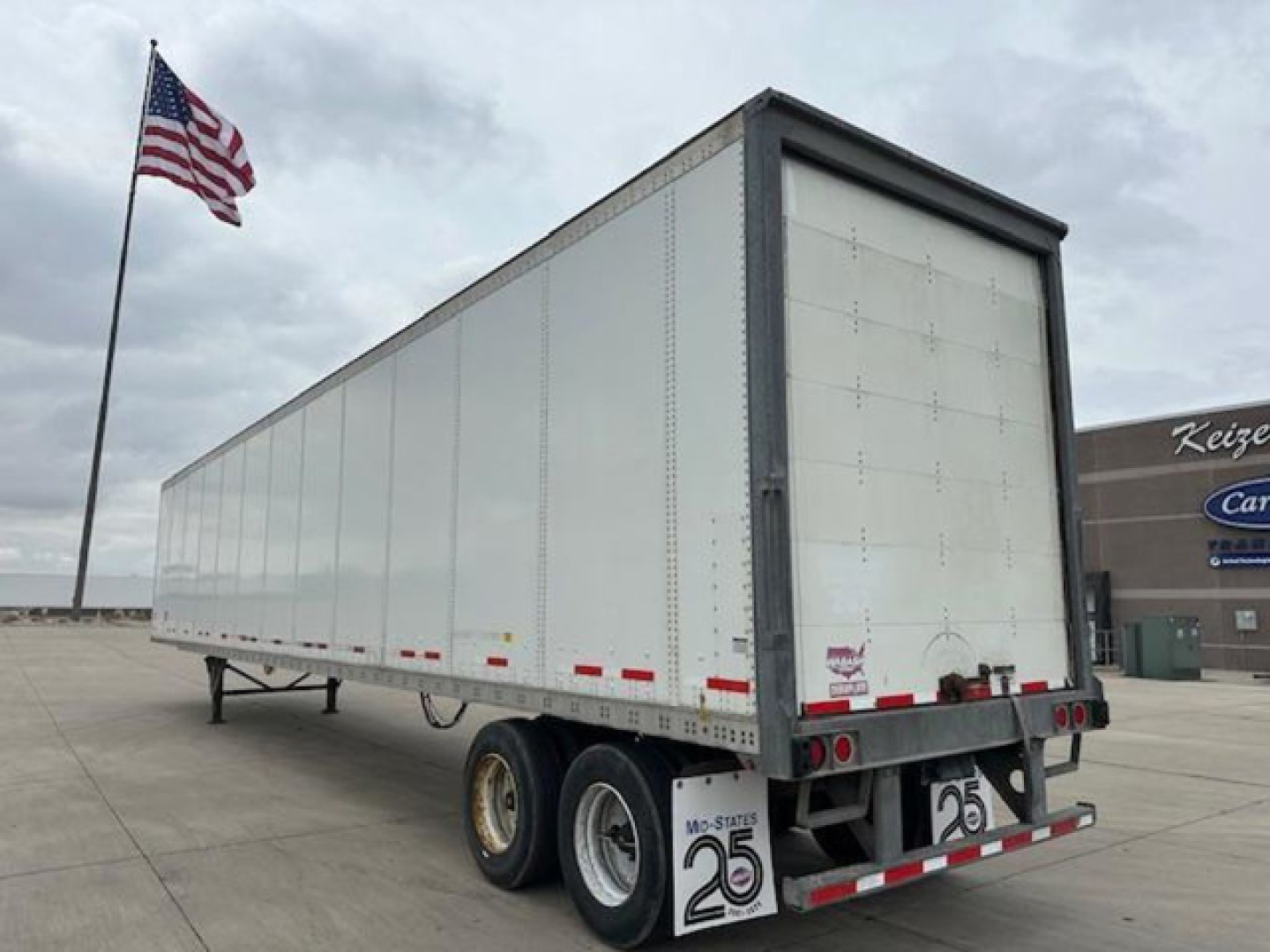 Trailer 53' Wabash van trailer showing rear end with dual axles and white exterior, American flag in background, stock numbers and markers on back.
